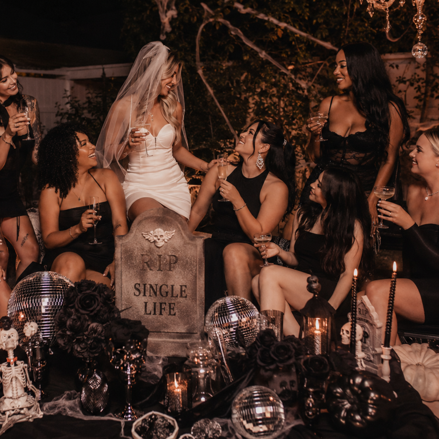 RIP single life bachelorette party with the bride and her friends celebrating outdoors beside gothic tombstones, candles, pumpkins, and spooky decor.